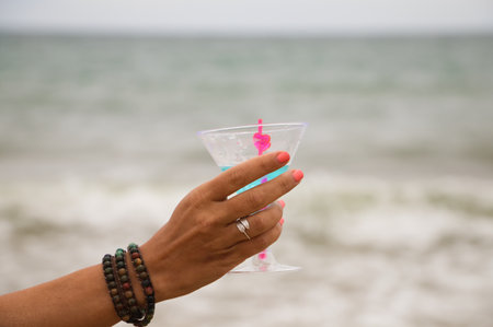 Detail of hand raising glass in the air with a blue drink. In the background you can see the sky and the sea in the horizon.の写真素材