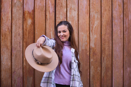 Young and beautiful Spanish woman from Seville with brown hair and plaid shirt holds a hat in her hand and offers it to the camera. The woman is happy and in the background a wooden door.の写真素材