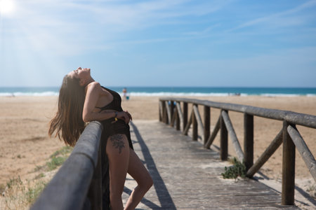 Beautiful young woman with long brown hair in swimming costume and black silk sarong leaning with her head tilted back on the railing of the walkway leading to the beach. The sea in the background.の写真素材