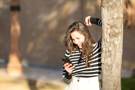 Young, beautiful woman with long brown hair wearing a black and white striped T-shirt leaning against a tree looking at her mobile phone. Concept of new technologies and telephonyの写真素材