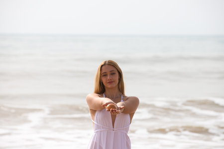 Beautiful young blonde woman in white dress holds a clam or shell in her hands and shows it to the front. The woman is happy and having fun. In the background the blue sea.の写真素材