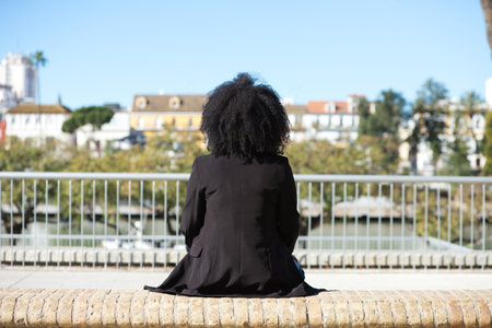 young, beautiful black woman with afro hair sitting on a bench facing the other way. Photo taken from behind.の写真素材