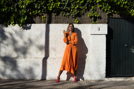 Pretty young blonde woman in orange dress poses for photos on white wall with ivy. The woman enjoys and has fun while doing different poses and expressionsの写真素材