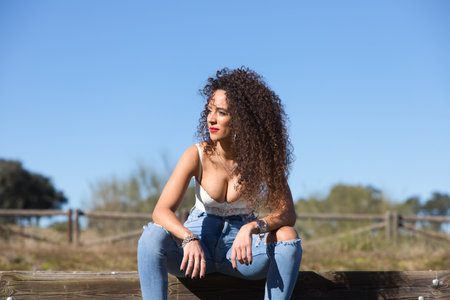 Spanish, brunette, curly-haired woman sits on a bench on the path leading to the forest. She is dressed in jeans and a white top and enjoys the sunny day.の写真素材