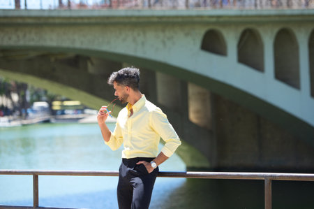 Handsome young man with beard and sunglasses in hand is leaning on the railing of the river guadalquivir in seville. On the other bank is the famous flamenco artists' quarter of Sevilleの写真素材