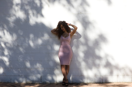 Beautiful brunette woman with curly hair leaning against a white wall between lights and shadows makes different body expressions, sadness, tiredness. The woman is dressed in an elegant pink and narrow dress.の写真素材