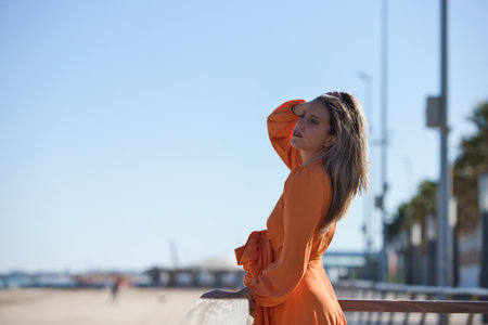 Young beautiful blonde woman in orange dress looks sadly leaning on the wooden railing on the beach. Blue sky in the background on the horizon. Fashion and beauty conceptの写真素材