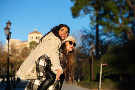 two young, beautiful Spanish women who are friends have fun playing on top of each other while they are walking and enjoying the sunny winter day. The women are wearing winter clothes.の写真素材