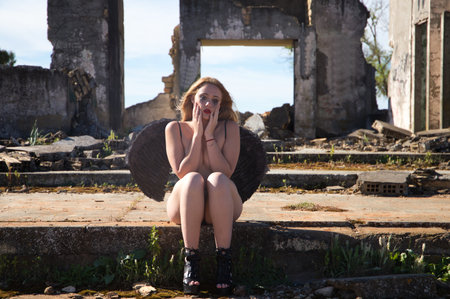 A black angel sitting with her hands on her face is crying in the ruins of a building after the apocalypse. Young, beautiful blonde woman with black wings and mascara on her face. She is sad.の写真素材