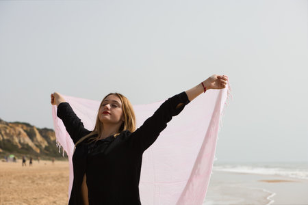 Young and beautiful blonde woman walks along the shore of the beach with a pink sarong in the wind, in the background the sea and the sky. Concept of freedom and holidays.の写真素材