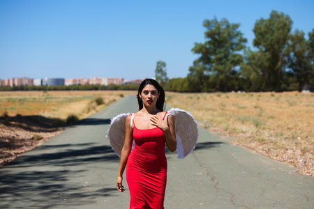 Beautiful young Latin woman in red silk dress and white wings walks along a tarmac road in the countryside outside the big city. The woman makes different poses and expressionsの写真素材