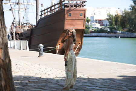 Young beautiful blonde woman dressed in embroidered trousers and crochet top by the river bank in Seville. In the background a replica of Christopher Columbus' boatの写真素材