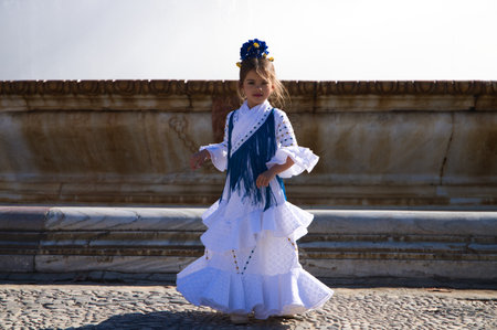 A pretty little girl dancing flamenco dressed in a white dress with ruffles and blue fringes in a famous square in Seville, Spain. The girl has a flower in her hair. In the background of a big fountainの写真素材