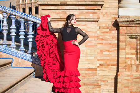 Beautiful woman dancing flamenco in a square in Seville, Spain. She is wearing a typical red and black dress and is leaning on a brick and tile wall and holding her red frilly dressの写真素材