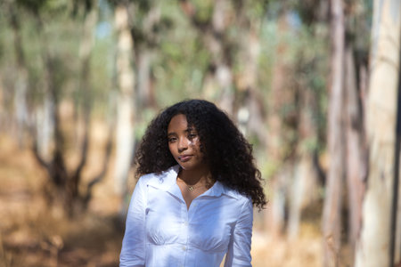 portrait of a young, beautiful, Latin, brunette woman with curly hair dressed in a white shirt looking at the camera among the eucalyptus trees in the forestの写真素材
