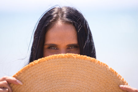 A pretty young brunette woman with green eyes covers half her face with a straw hat, revealing her eyes. In the background the horizon with the sea and the blue sky. Beauty and fashion conceptの写真素材