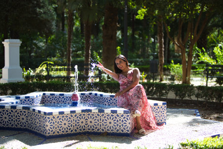 beautiful young brunette woman in a long flower patterned dress is sitting in a star-shaped fountain, the woman splashes water with her hand and is having fun. Travel and holiday conceptの写真素材