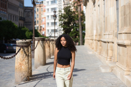 beautiful young latin woman with black eyes and dark, long curly hair walks through the center of Seville next to the archive of the india and some 15th century stone columns. she is in Andalusiaの写真素材