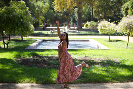 young and beautiful brunette woman in long dress with floral print poses for photo and lifts one arm and one leg and enjoys herself. The woman enjoys the sunny day in the cityの写真素材