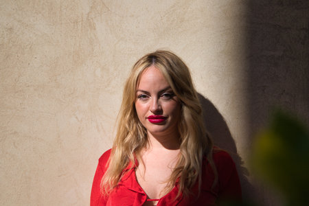 blonde woman, young and beautiful, make-up, dressed in red and elegant, looks at the camera in a sensual and serious way. In the background a pink wall. Beauty and fashion conceptの写真素材