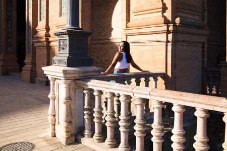 young and pretty brunette woman dressed in white top and jeans is happy leaning on the railing in the square of Spain in Seville, she looks back and takes the photoの写真素材