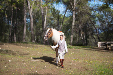 Equestrian woman, young and beautiful, holds the reins and walks through a pine forest with her purebred brown horse with blond mane. The woman is happy. Concept of love for the horseの写真素材