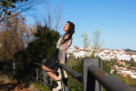 Latin woman, young and brunette sitting on a fence at a viewpoint in a village in andalusia. In the background the village of white houses and the church.の写真素材