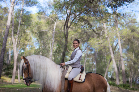 Equestrian woman, riding and holding the reins of her purebred brown horse with blonde mane and tail. The woman gallops through the forest. Concept of love for the horseの写真素材