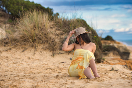 Young, beautiful brunette woman in yellow dress is sitting on the fine white sand on the cliff. The girl is relaxed and happy, holding her straw hat in her hand and looking downの写真素材