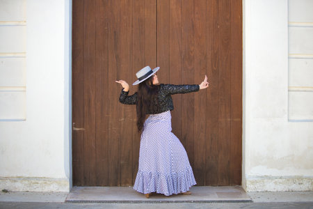 Latin woman, young and beautiful dancing flamenco in front of a wooden door in a typical street of Andalusia. The woman is dressed in a white ruffled dress with black polka dots, a hat and a jacketの写真素材