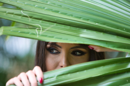 Pretty young brunette woman clutches a palm leaf in her hands and looks through it. The woman is very expressive with the big black eyes of a Spanish woman. Beauty and fashionの写真素材