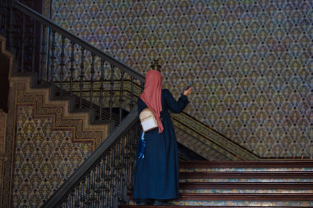 a muslim woman with red turban on her head and black tunic walks on the stairs of the famous square of spain in seville, andalusia, the woman is recording with her mobile phone the monumentの写真素材