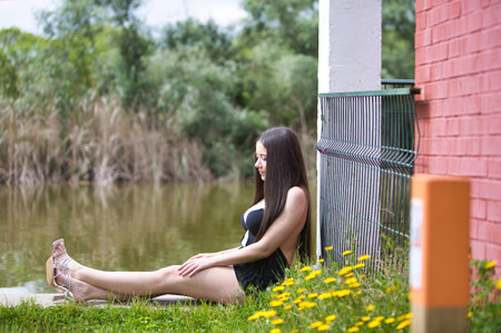 Young, dark and beautiful woman dressed in black is sitting by the lake between yellow flowers and grass on the ground. In the background of a lake.の写真素材