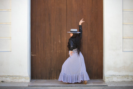 Latin woman, young and beautiful dancing flamenco in front of a wooden door in a typical street of Andalusia. The woman is dressed in a white ruffled dress with black polka dots, a hat and a jacketの写真素材