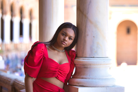 African woman, with long brown hair, young and beautiful, dressed in an elegant red dress, rests her arm on a white marble column in the square Spain in Seville, Andalusia. The woman is seriousの写真素材