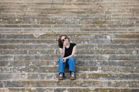young and beautiful blonde woman, tattooed and wearing jeans, black shirt and sunglasses, sitting on the stairs doing different postures and expressionsの写真素材