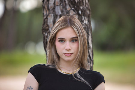 Portrait of beautiful young blonde woman in black shirt leaning against a tree trunk in the forest. The woman is serious and staringの写真素材