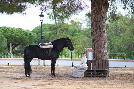 Beautiful young woman sitting in the shade of a pine tree next to her black purebred Spanish horse in Andalusia, Spain. The woman is dressed in typical horsewoman costumeの写真素材