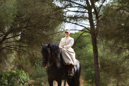 Young and beautiful woman riding a purebred Spanish black horse in a pine forest. The woman is dressed in typical horsewoman costume. In the background the lush vegetationの写真素材
