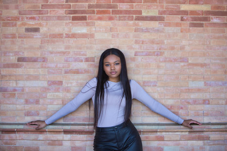African woman, young and beautiful dressed in beige top and black leather shorts leaning on a red brick wall with her arms outstretchedの写真素材