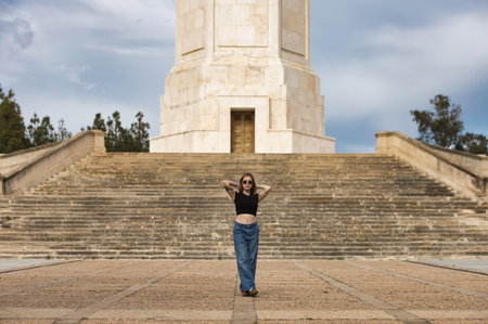 young and beautiful blonde woman, tattooed and wearing jeans, black shirt and sunglasses, poses doing different postures and expressions in front of some stairs going up to a towerの写真素材