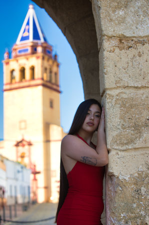 A beautiful teenage girl with long dark hair dressed in red leaning under a Roman archway on a street in a typical Andalusia town. In the background is a church towerの写真素材