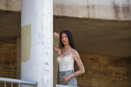 Young and pretty brunette with tattoos on her arms and dressed in casual clothes leaning on a concrete column. The woman is wearing makeupの写真素材