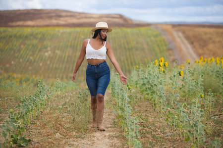 A beautiful black Latin farmer wearing a white top, jeans and a straw hat walks around observing the sunflower plantation. The woman is happy and smiling on this sunny summer day in the countrysideの写真素材