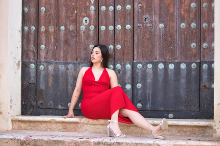 A pretty teenage girl with long dark hair, dressed in red and wearing high heels, sitting on the steps of a church and leaning against the wooden doorの写真素材