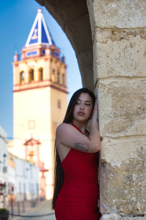 A beautiful teenage girl with long dark hair dressed in red leaning under a Roman archway on a street in a typical Andalusia town. In the background is a church towerの写真素材