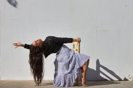 Latin woman, young and beautiful, dancing flamenco sitting on an old wooden chair and throwing her head back. The woman is dressed in a white ruffled dress with black polka dots and a black jacketの写真素材