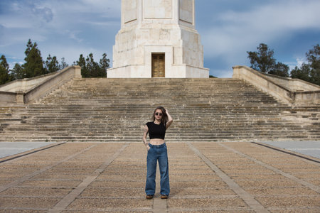 young and beautiful blonde woman, tattooed and wearing jeans, black shirt and sunglasses, poses doing different postures and expressions in front of some stairs going up to a towerの写真素材