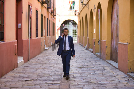 A young, handsome, dark-haired businessman wearing a suit and tie and black lace-up shoes walks along talking on the phone. The man is the director of a multinational companyの写真素材