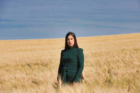 A beautiful, dark-haired teenage Latin girl dressed in green stands among a dry, yellow wheat field on a summer day. In the background, the blue skyの写真素材